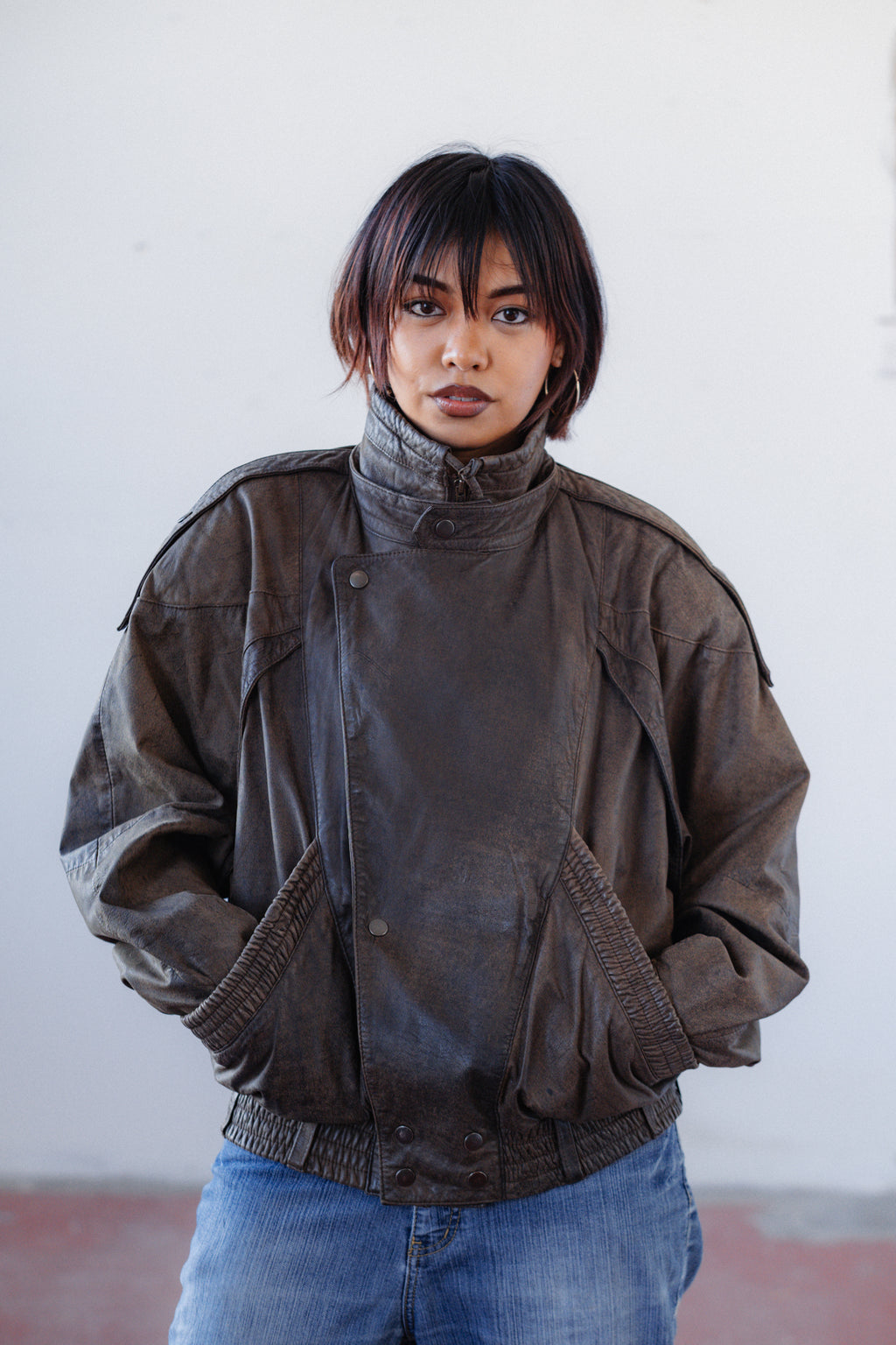 A model wearing a dark brown, distressed military-style jacket with a high collar and front flap pockets, paired with blue jeans, standing against a plain off-white wall.