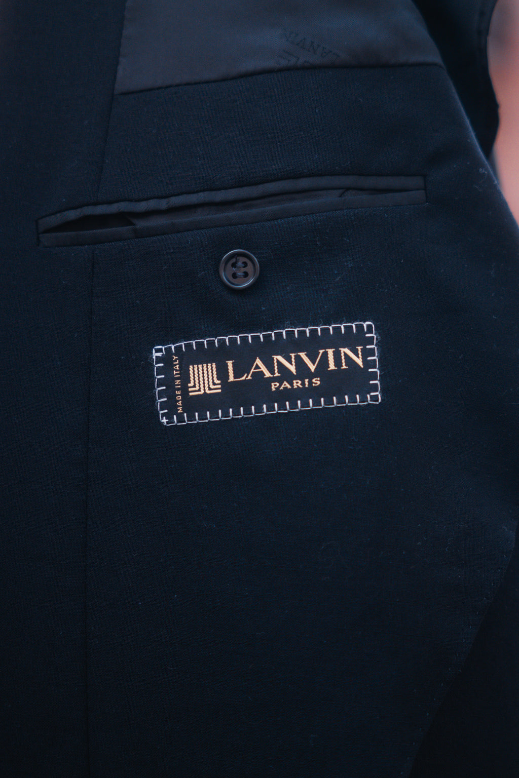 Close-up of a dark navy or black tailored jacket or vest with a visible Lanvin Paris brand patch stitched onto the front, showing part of a button and pocket detail.