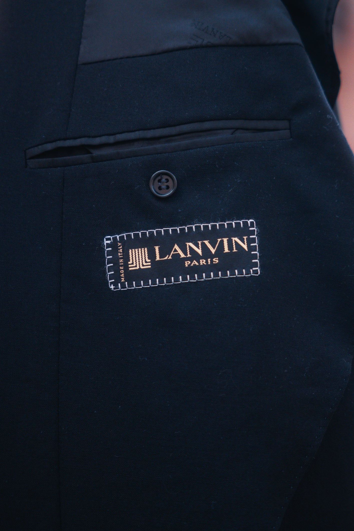 Close-up of a dark navy or black tailored jacket or vest with a visible Lanvin Paris brand patch stitched onto the front, showing part of a button and pocket detail.