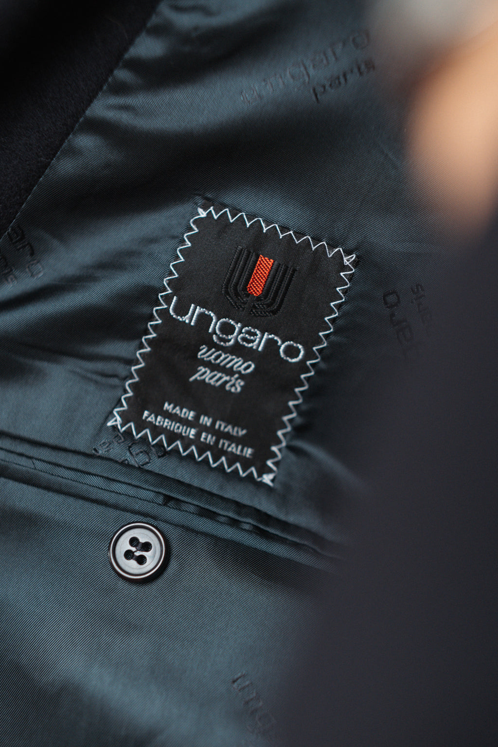 Close-up of a dark teal or slate-colored jacket lining with a sewn black brand label reading 'ungaro uomo paris' and a small red accent, near a button.