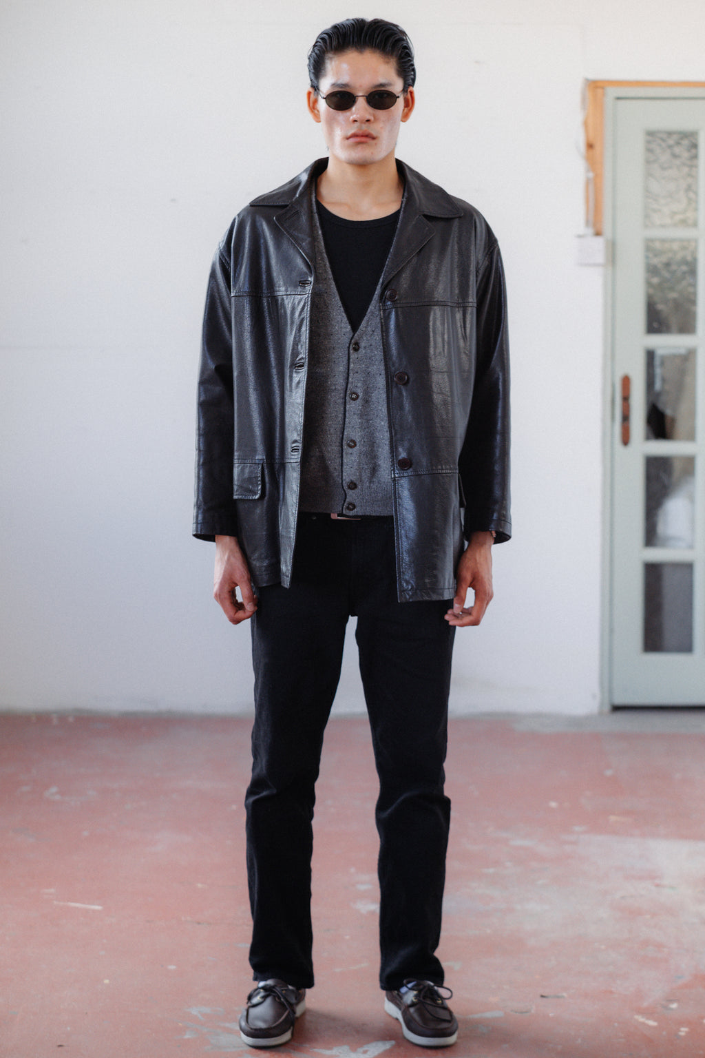 A man wearing a black leather jacket over a gray button-front vest, black shirt, black pants, and black sneakers, standing in a minimalist room with a white wall and a reddish-brown floor.