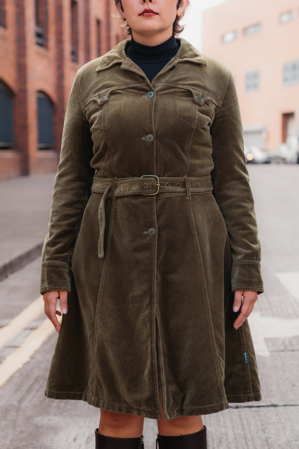 A woman wearing an olive green corduroy trench coat with a matching belt, styled over a black turtleneck and black boots, standing on a city street.