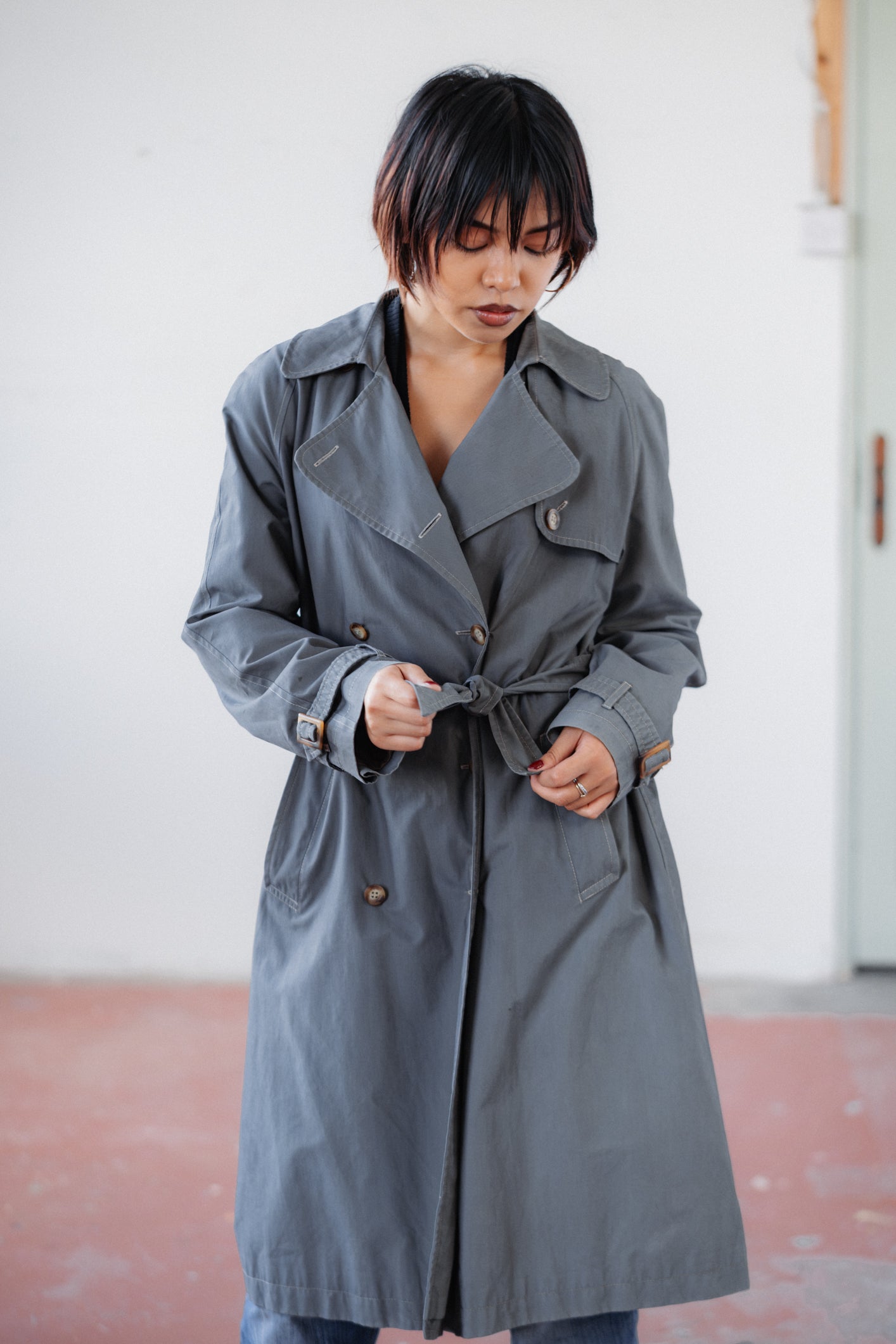 A person wearing a long gray double-breasted trench coat with a self-tie belt, standing in a minimalist indoor setting with a white wall and reddish-brown floor.
