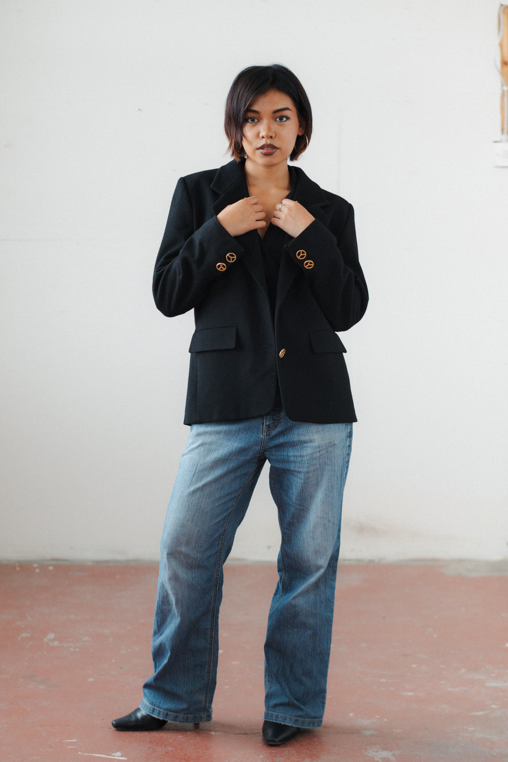 A woman stands in a studio wearing a black blazer with gold buttons, blue jeans, and black shoes against a plain white wall and reddish-brown floor.