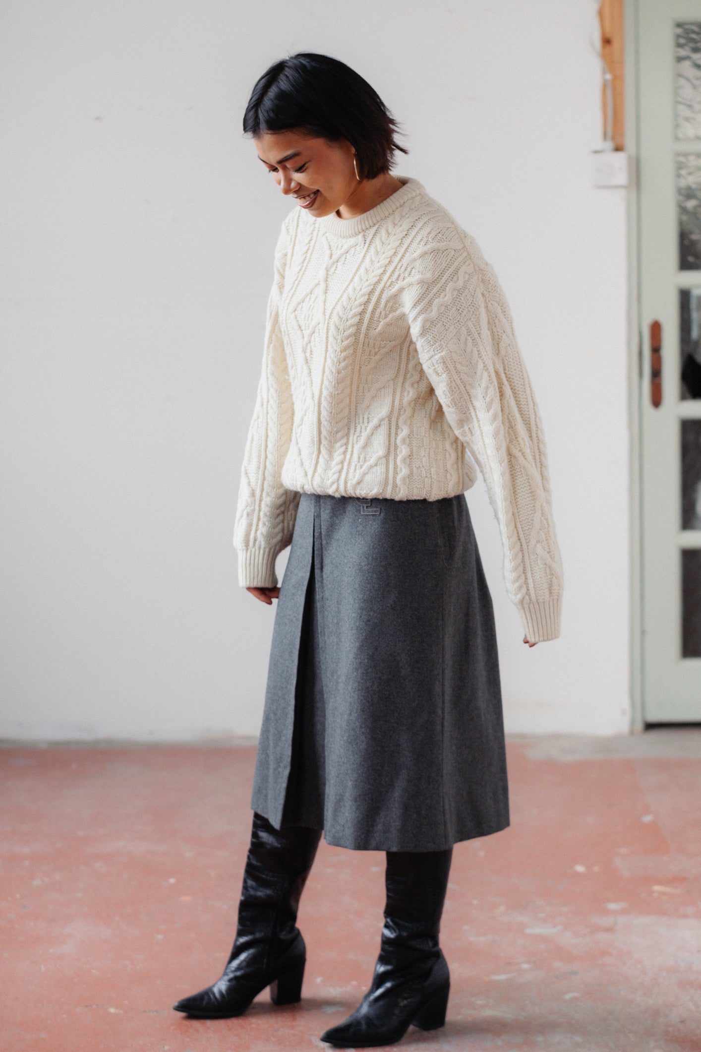 A woman wearing a cream cable-knit sweater, a dark gray midi skirt, and black knee-high boots, standing in a minimalist room with a white wall and reddish-brown floor.