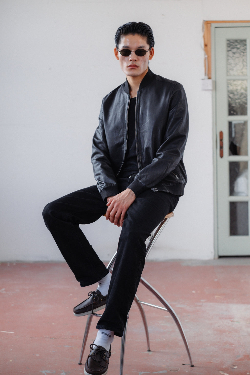 A man wearing a black leather jacket, black pants, white socks, and black loafers sits on a metal stool in a minimalist indoor setting with a white wall and a light-colored door.