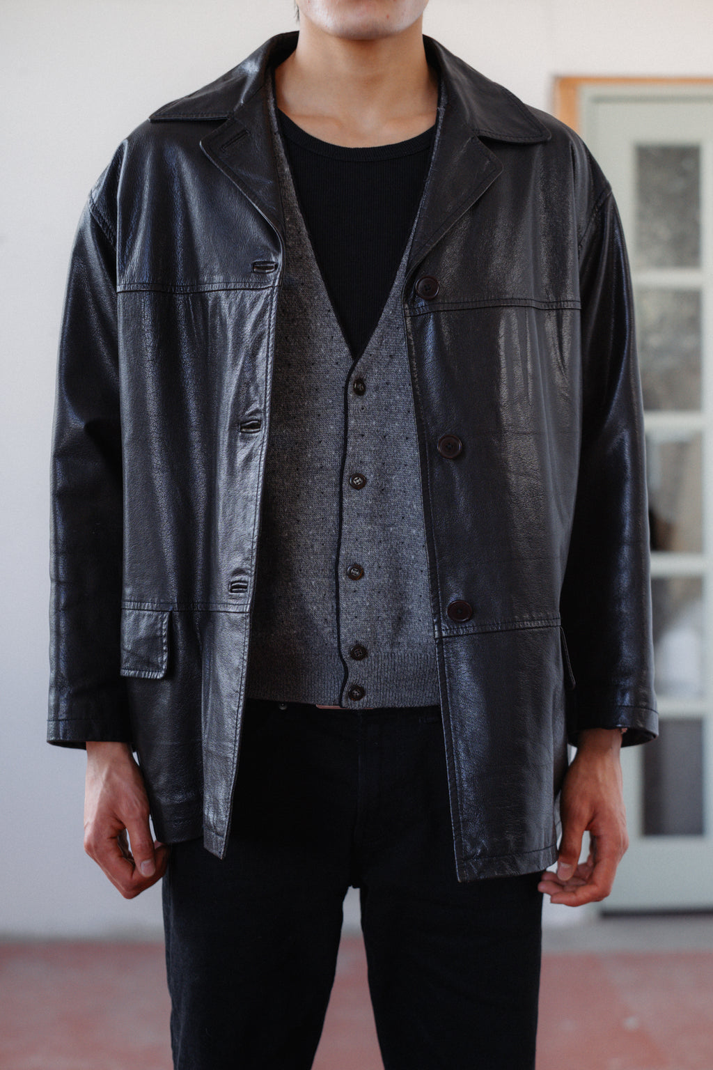 A person wearing a black leather jacket over a gray button-front vest, black shirt, and black pants, standing indoors against a light wall and window frame.
