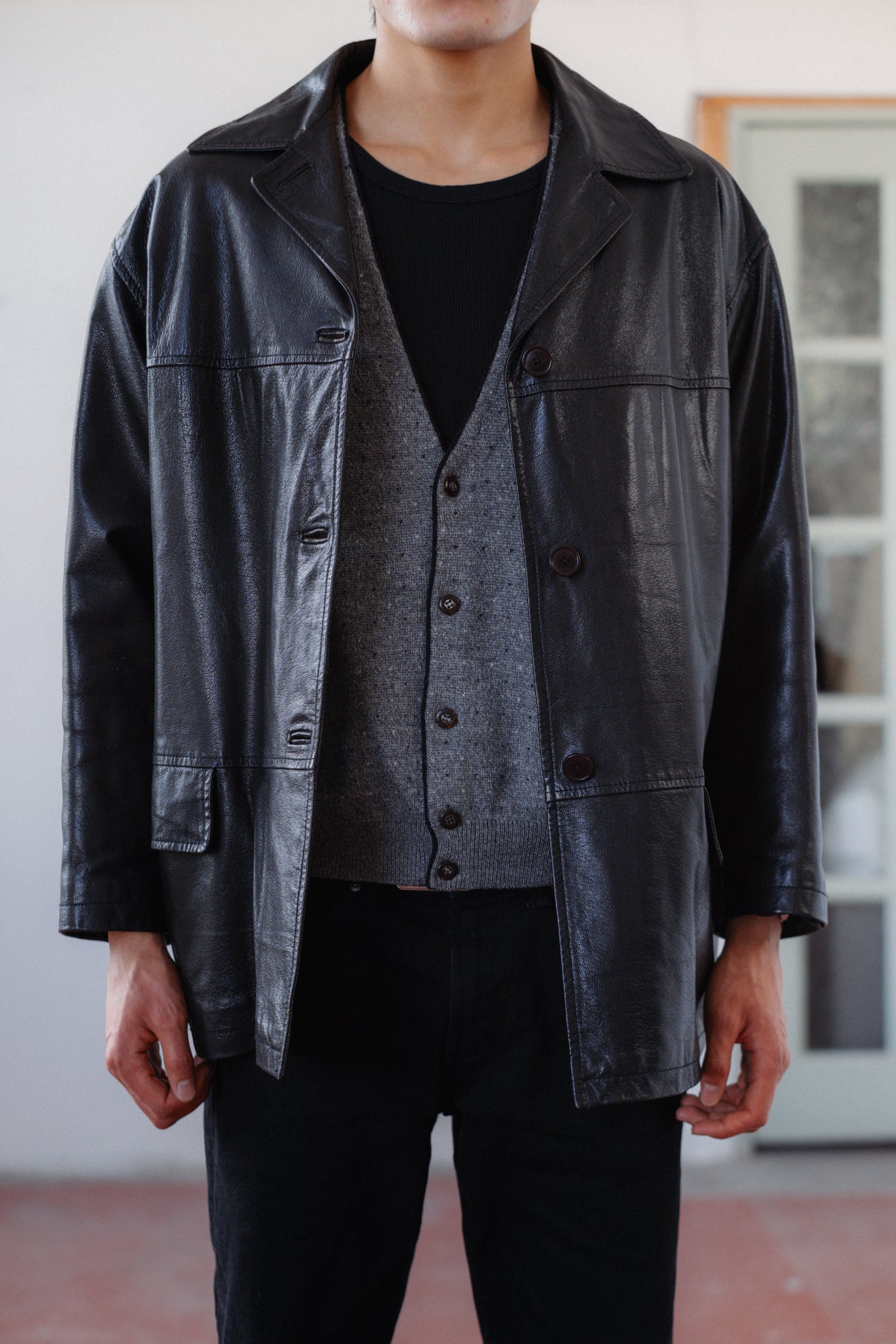 A person wearing a black leather jacket over a gray button-front vest, black shirt, and black pants, standing indoors against a light wall and window frame.